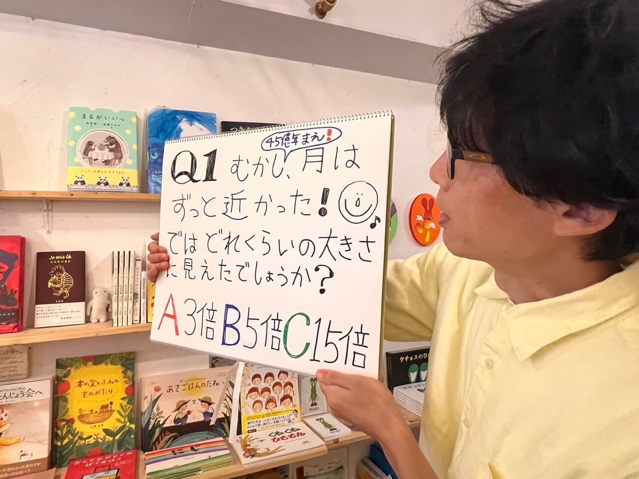 「8月24日のニジノ絵本屋１号店でのイベントの様子⑤／バスボムが乾くのを待っている間にお楽しみタイム♪ カワチ・レンさんによる、おつきさまクイズ。おつきさま色のシャツ姿の方がカワチ・レンさんです」画像